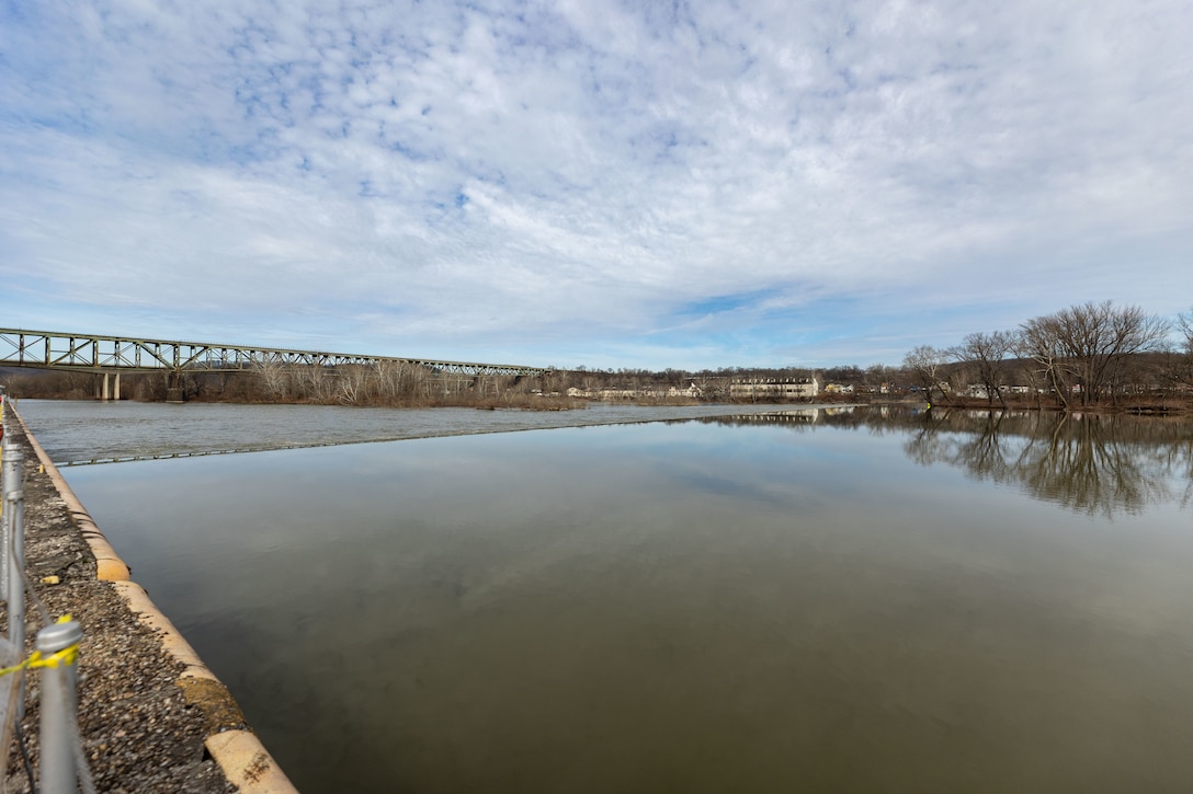 A wide, panoramic view of a calm river with a bridge in the distant background under a cloudy sky.
