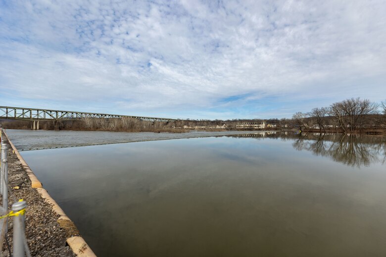 A wide, panoramic view of a calm river with a bridge in the distant background under a cloudy sky.
