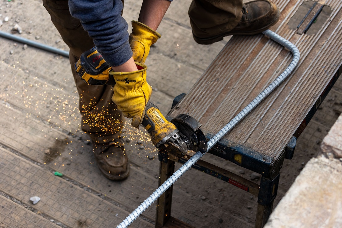 An overhead view of a person wearing yellow gloves using a power grinder to cut a metal bar, with sparks flying.