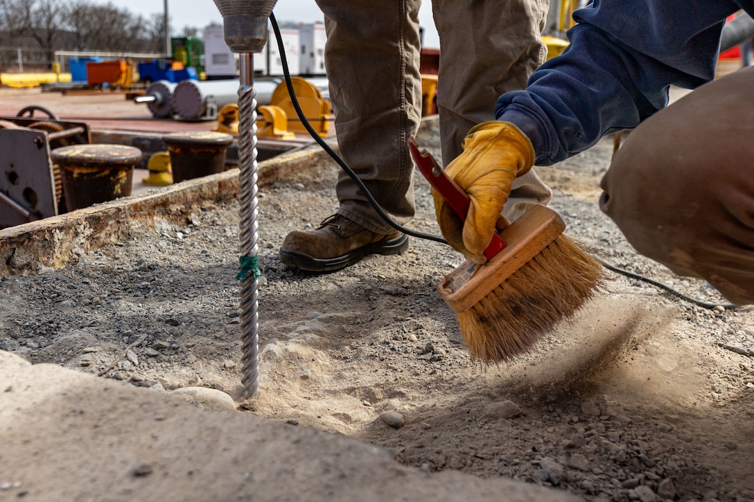 A close-up of a worker using a brush to clear dust away from a large drill bit drilling into concrete.