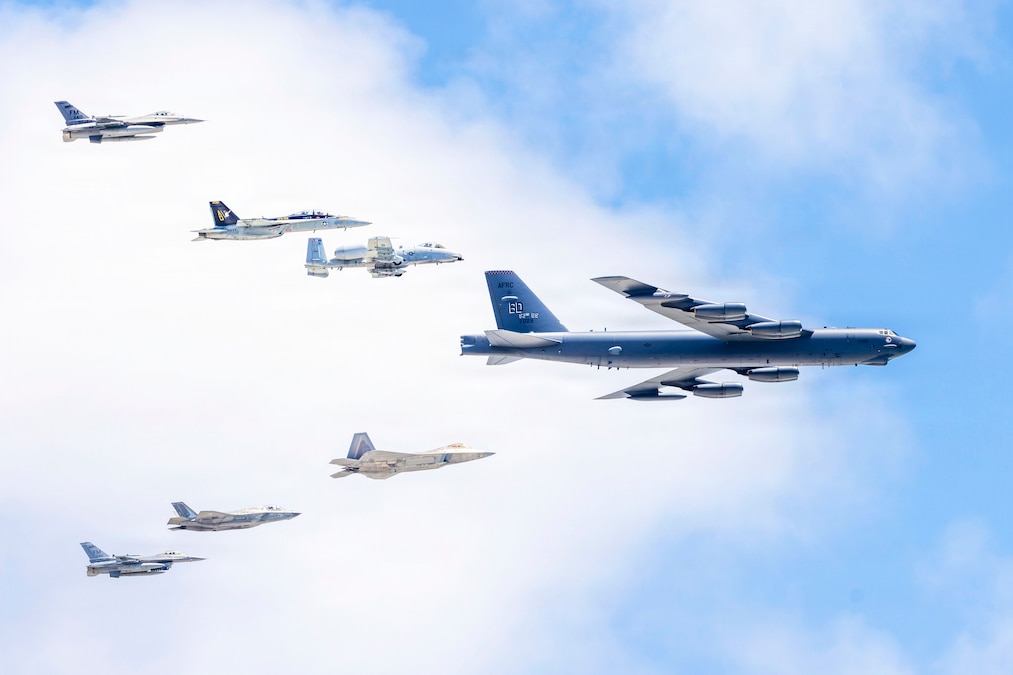 An aircraft leads six other aircraft traveling in a triangular formation in a blue sky.