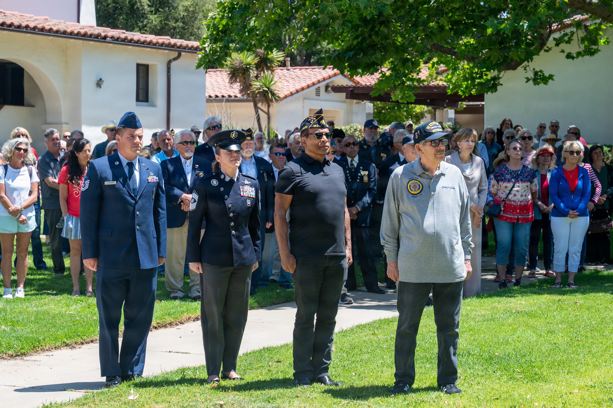 People stand on grass outside during a ceremony. Four people stand in front of a crowd at the position of attention.