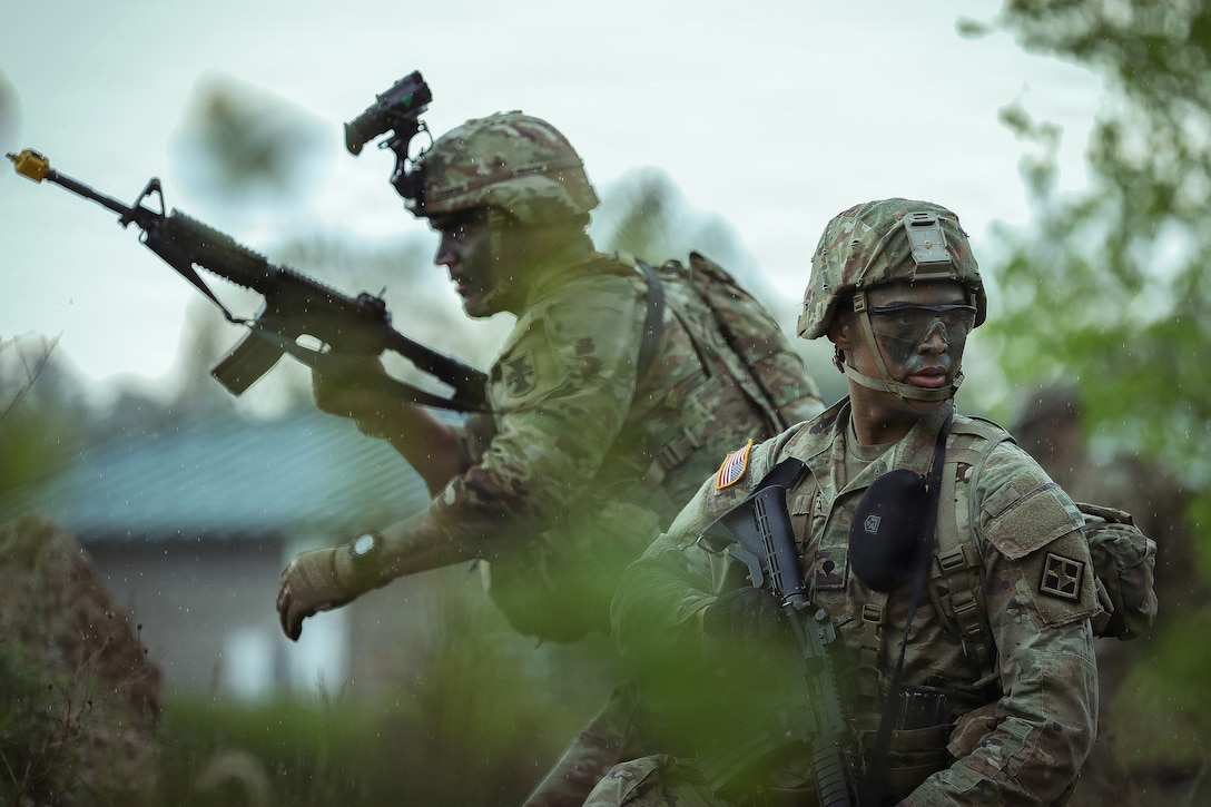 Two soldiers in camouflage uniforms hold machine guns and look in opposite directions outside. They are surrounded by tall grass and trees are in the background.