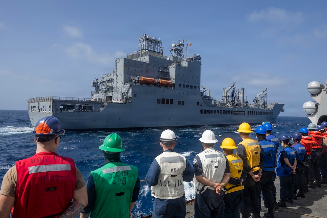 People in hard hats and vests over their clothes stand in a row by the edge of a ship’s deck looking over at another ship that is in the water nearby.