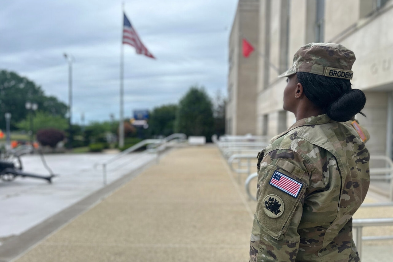 A woman wearing a camouflage military uniform salutes the American flag outside.