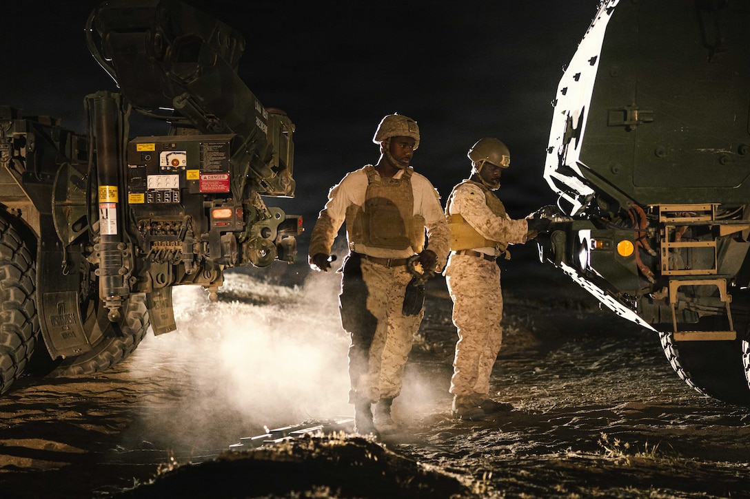 Two Marines stand in between two vehicles at night, with smoke on the ground. One Marine is holding and looking at something on the back of one of the vehicles.