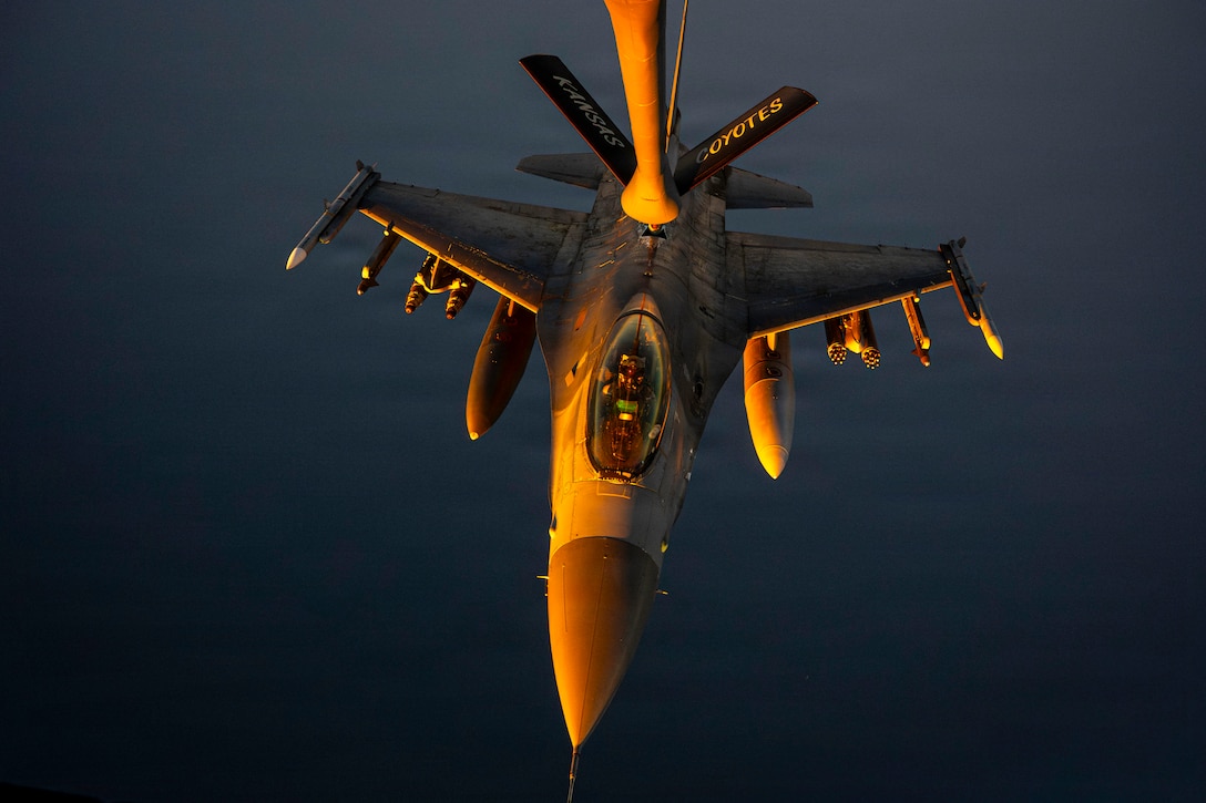 A fighter jet is in the air, with a dark sky in the background part of a refueling aircraft above it.