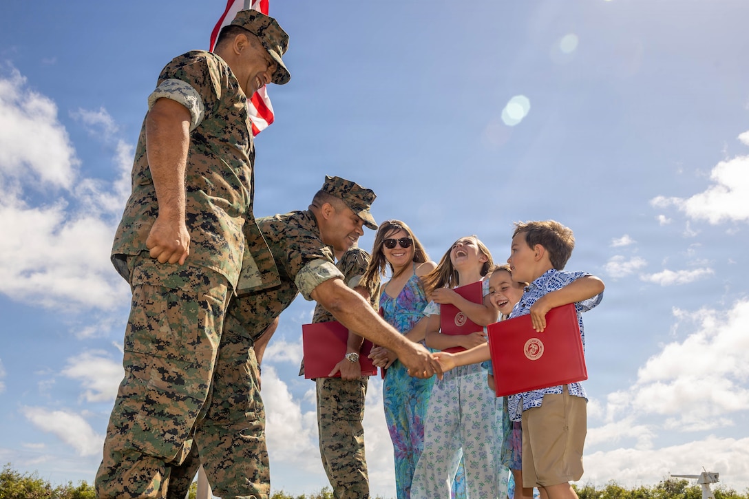 A Marine shakes hands with a child holding a red folder as another Marine, a civilian, and several smiling kids look on.