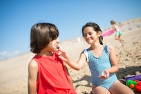 Two children sit on a beach while one applies sunscreen on the other's face.