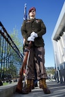 An Army Soldier wearing dress greens holds a rifle at order arms.