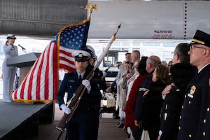 A man in a formal military uniform holds a rifle while leading a line of people in similar attire; one is carrying an American flag. To the right, audience members stand with their hands over their hearts.