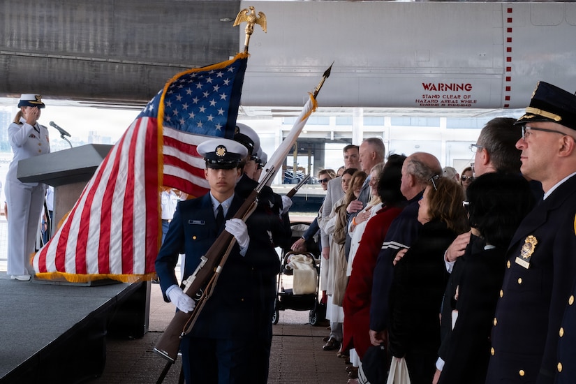 A man in a formal military uniform holds a rifle while leading a line of people in similar attire; one is carrying an American flag. To the right, audience members stand with their hands over their hearts.