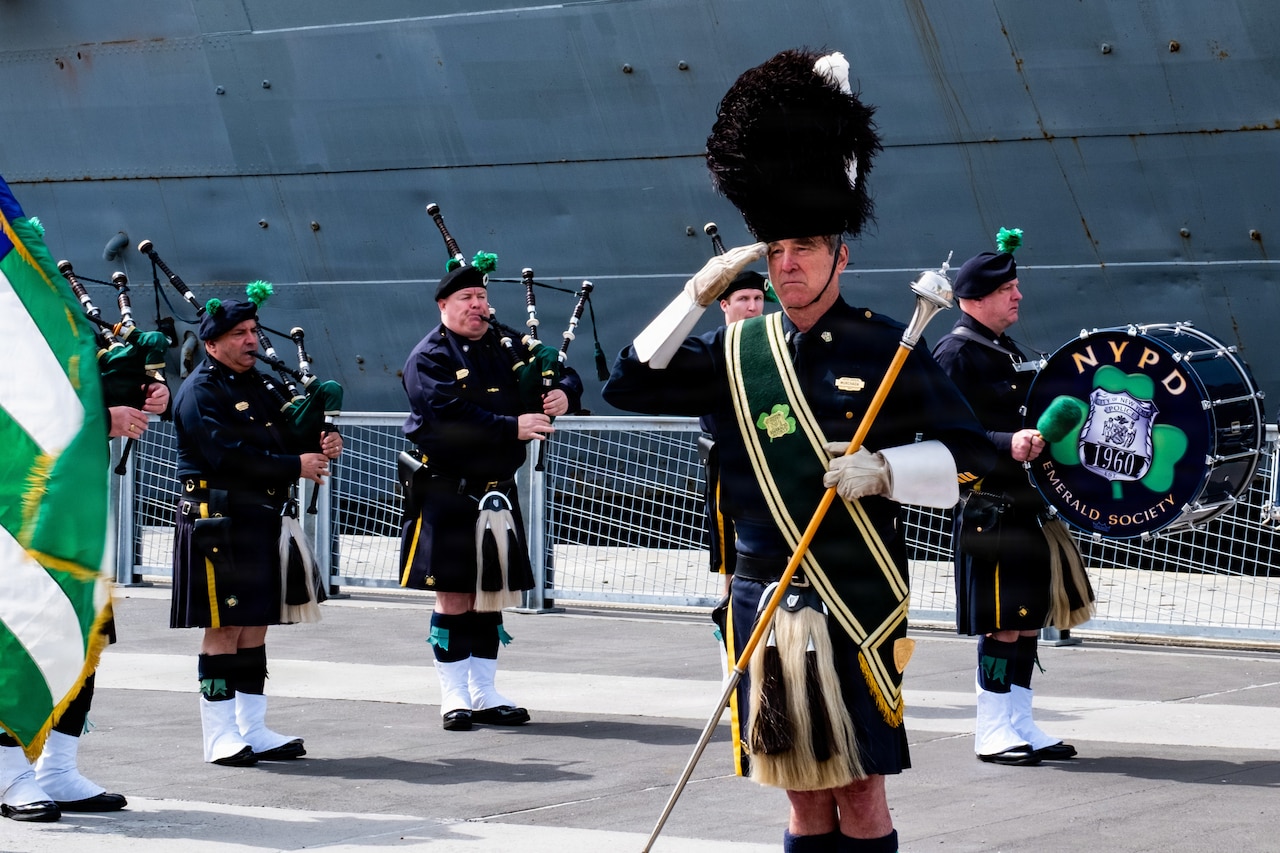 People in Irish attire play the bagpipes while one person salutes and another plays a large drum that reads, "NYPD - Emerald Society."