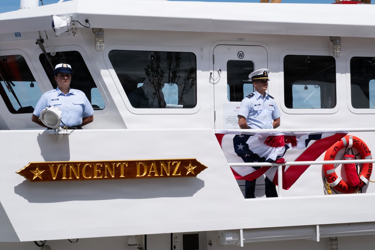 Two people in formal military uniforms stand on a ship; a sign on the ship reads, "Vincent Danz."