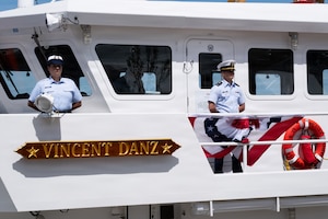 Two people in formal military uniforms stand on a ship; a sign on the ship reads, "Vincent Danz."