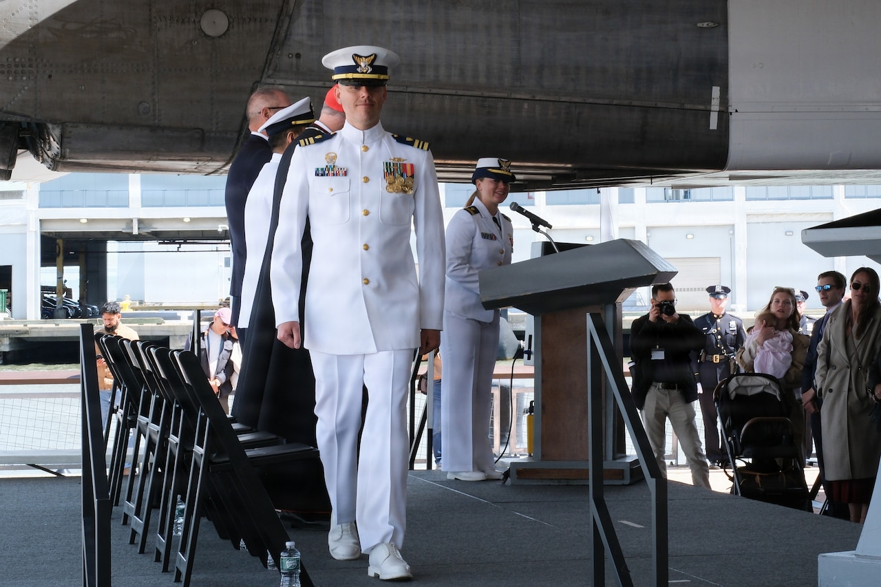 A man in a formal military uniform walks off a stage while people in similar attire observe.