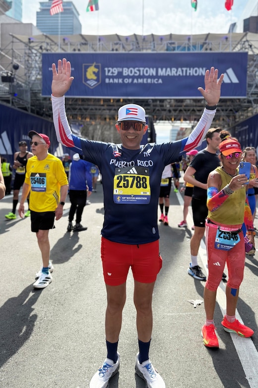 An airman wearing running gear holds both arms in the air on a road during daytime, with other people in athletic attire standing in the background.