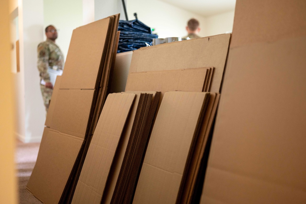 A close-up view of flat boxes leaning against a wall; a man wearing a camouflage military uniform is in the background.