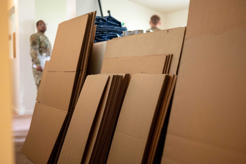 A close-up view of flat boxes leaning against a wall; a man wearing a camouflage military uniform is in the background.