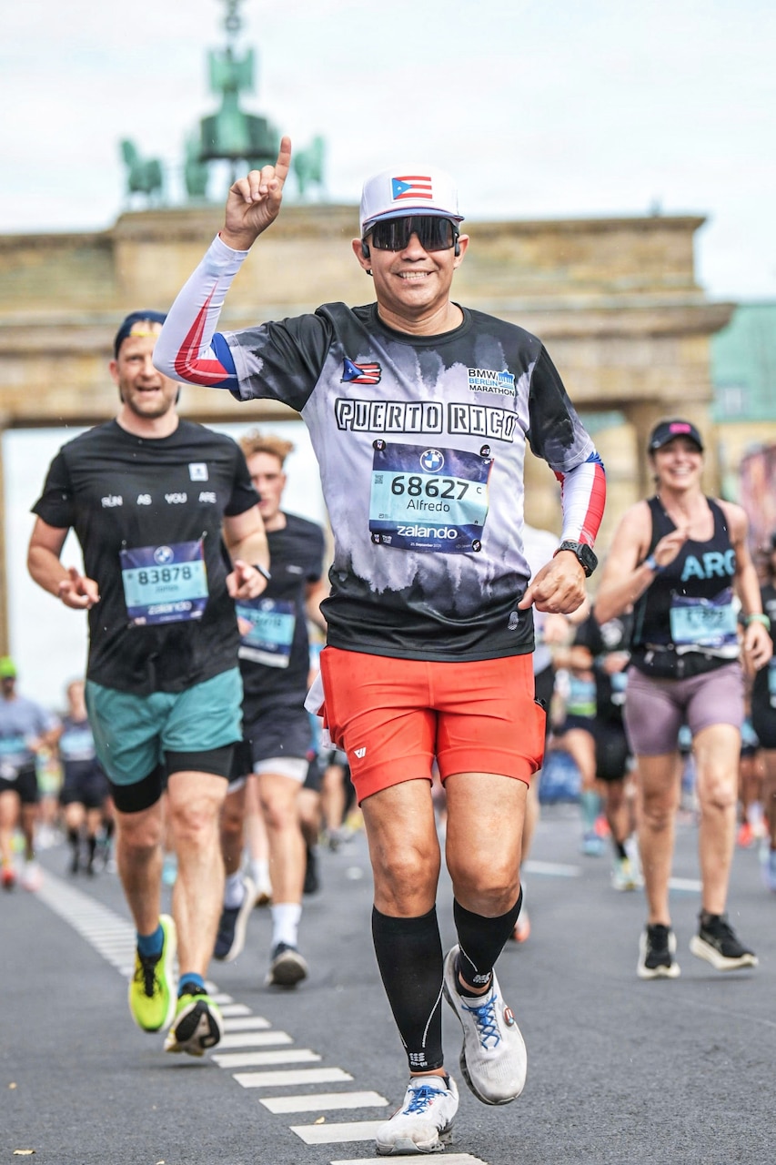 An airman wearing running gear holds one hand in the air and smiles as he jogs on a road during daytime, with other people in athletic attire also running in the background.