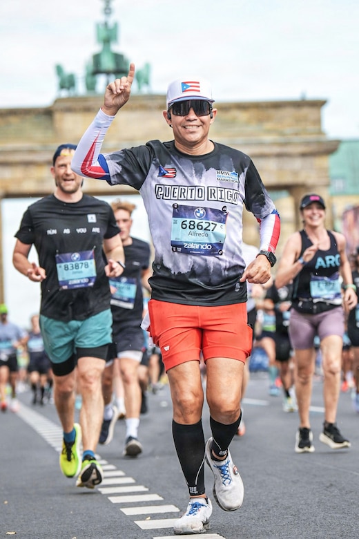 An airman wearing running gear holds one hand in the air and smiles as he jogs on a road during daytime, with other people in athletic attire also running in the background.