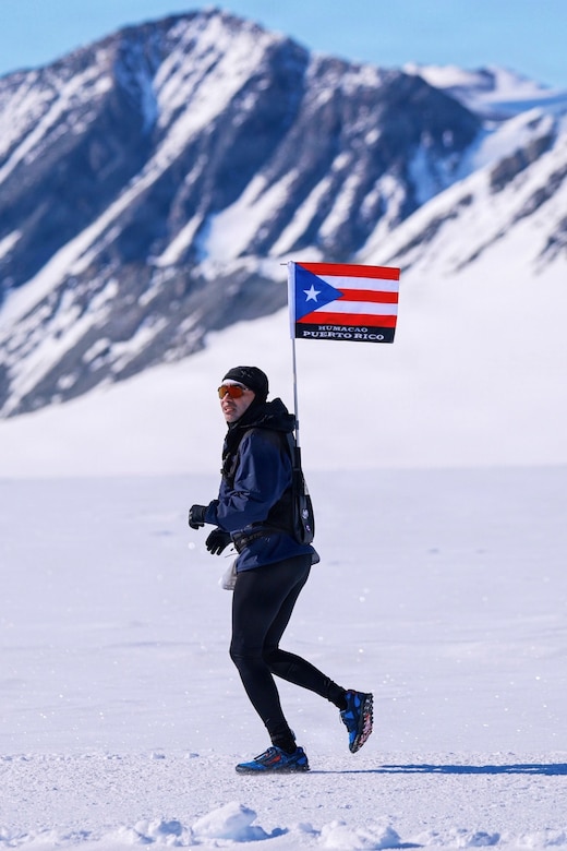 An airman in winter athletic gear and carrying Puerto Rico's flag on his back runs on ice during daytime; a snowy mountain is in the background.