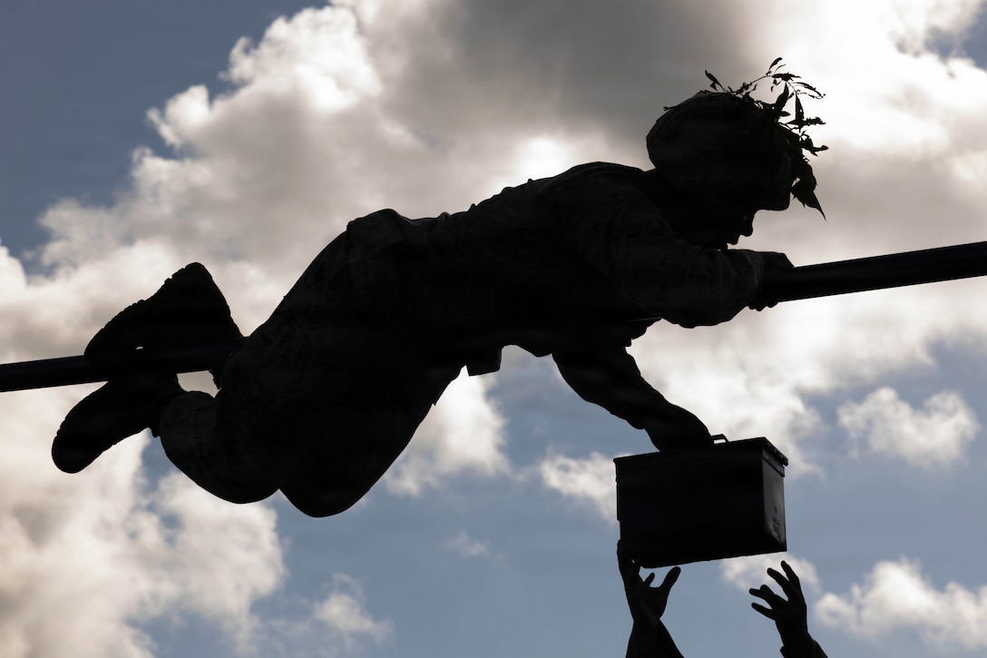 A Marine Corps recruit climbs across a pole in silhouette against a blue sky while holding a box as hands in the foreground reach for it.