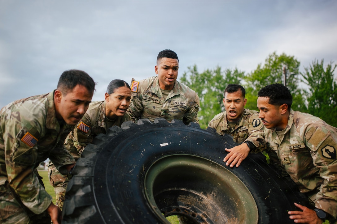 Five soldiers work together to flip a large tire in a field under a gloomy sky.