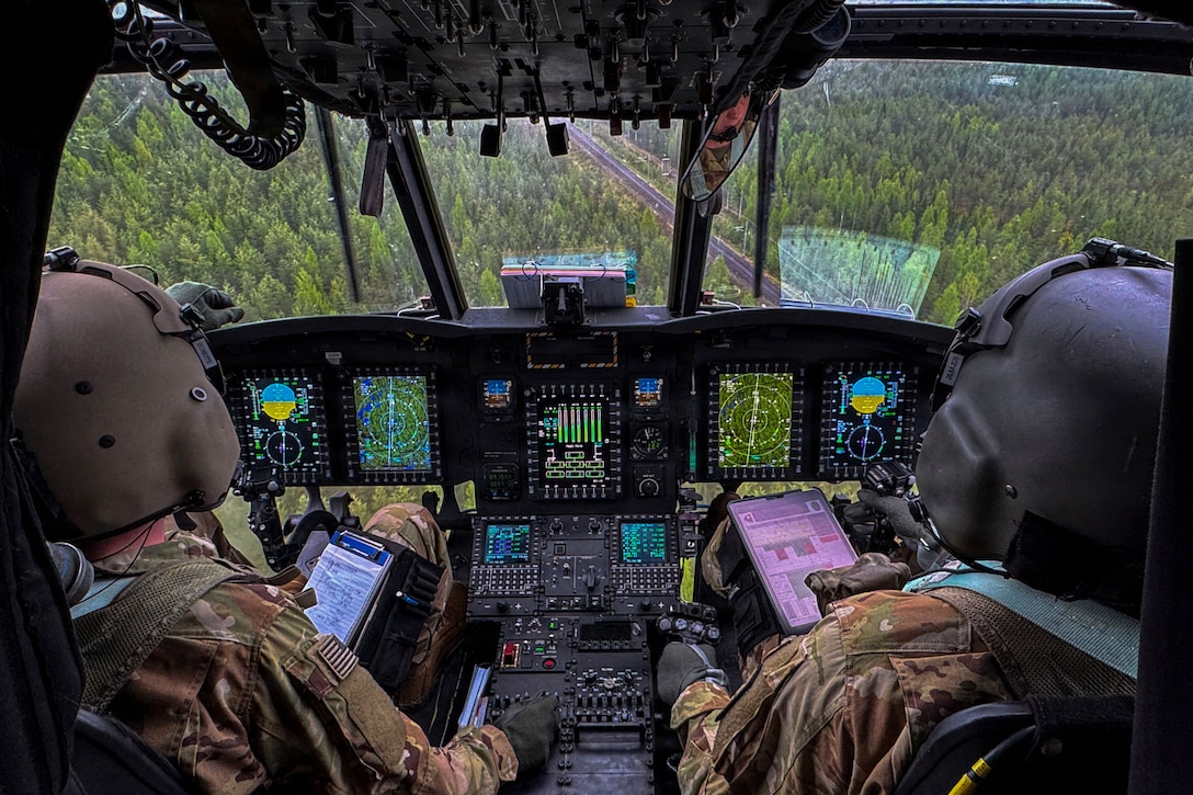 Two soldiers wearing protective gear sit in front of a control panel in a cockpit as dozens of trees are seen through the windshield.