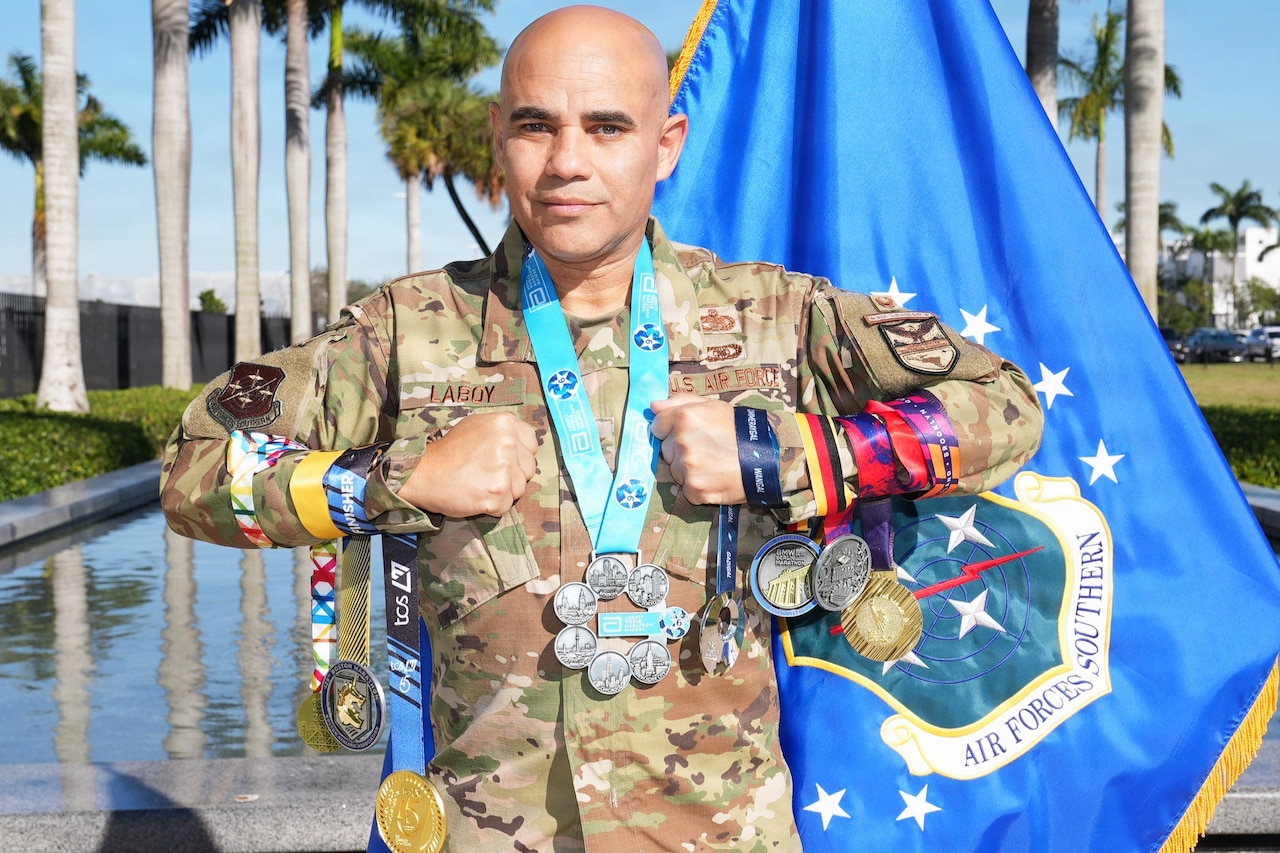 An airman wearing a camouflage uniform stands outdoors during daytime and holds seven medals wrapped around his arms; another large medal hangs around his neck, with a flag, tall trees and a reflecting pond in the background.