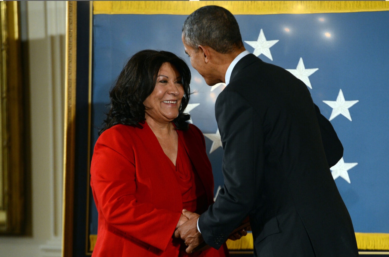 A man and a woman in business attire shake hands and smile in front of a blue square with stars.
