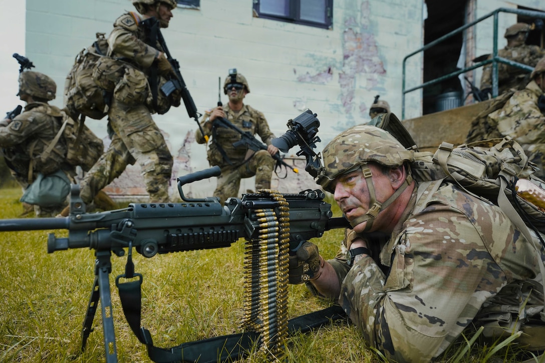 A soldier on the ground and looking straight ahead holds onto a gun positioned in front of him. Other soldiers and a white building are in the background.