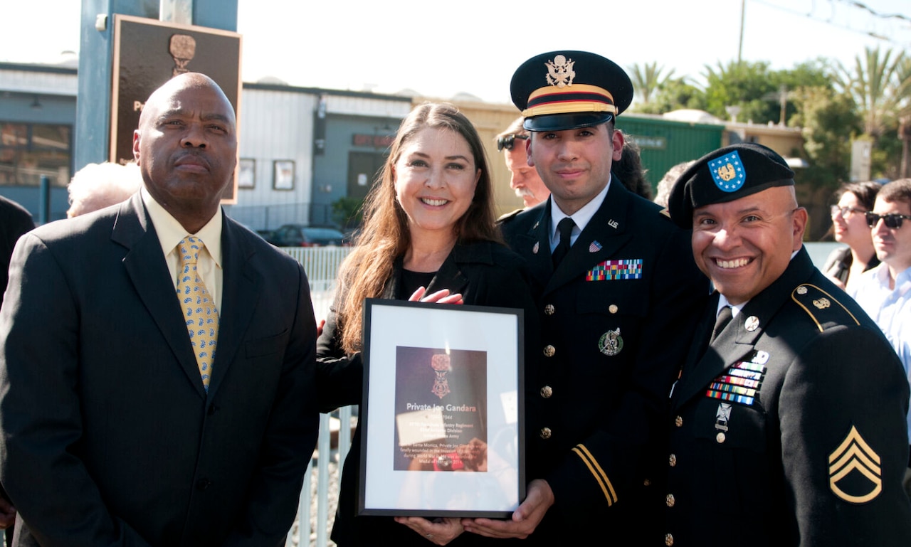 A woman in business attire holds a framed photo and poses outside with three men, two in formal military uniforms and one in business attire.