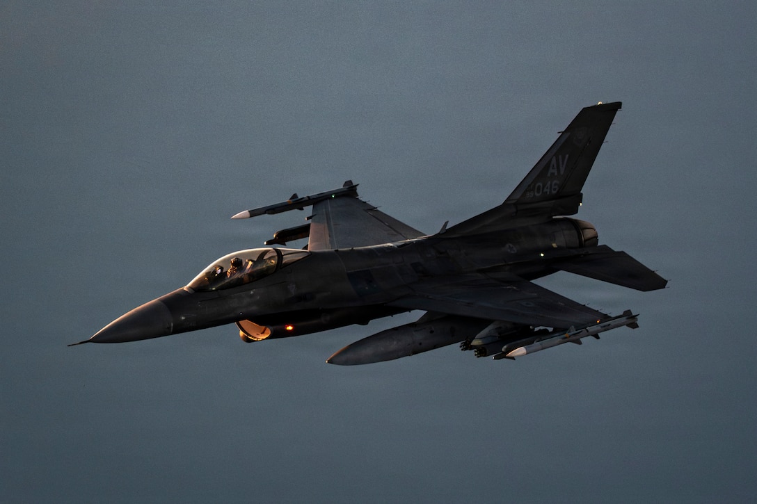 A pilot flies a fighter jet, with a muted gray-blue sky in the background.