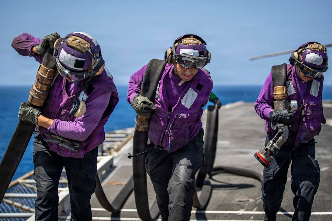 Three people in black pants and purple tops drag a black fuel hose on the deck of a ship, with blue water and the horizon in the background.