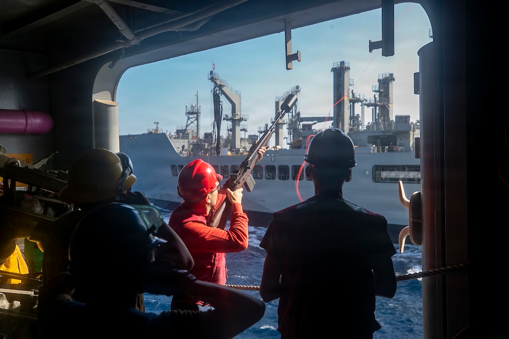 A sailor wearing a red helmet stands on the side of a ship and launches a red nylon cord from a modified rifle to another ship, with other people in silhouette also shown.