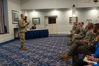 A man in camouflage stands on the left holding a microphone while speaking to a seated audience on the right.