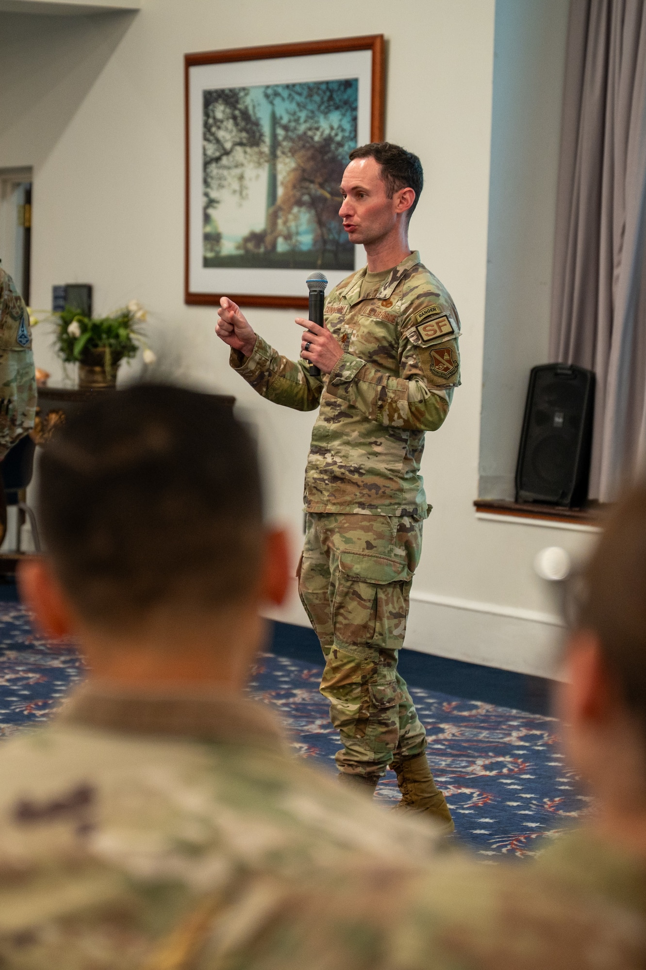 A man in camouflage speaks into a handheld microphone, standing between two seated people in the foreground.