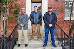Photograph of three men from NSWC Indian Head's COMSEC team standing on steps in front of a brick building.