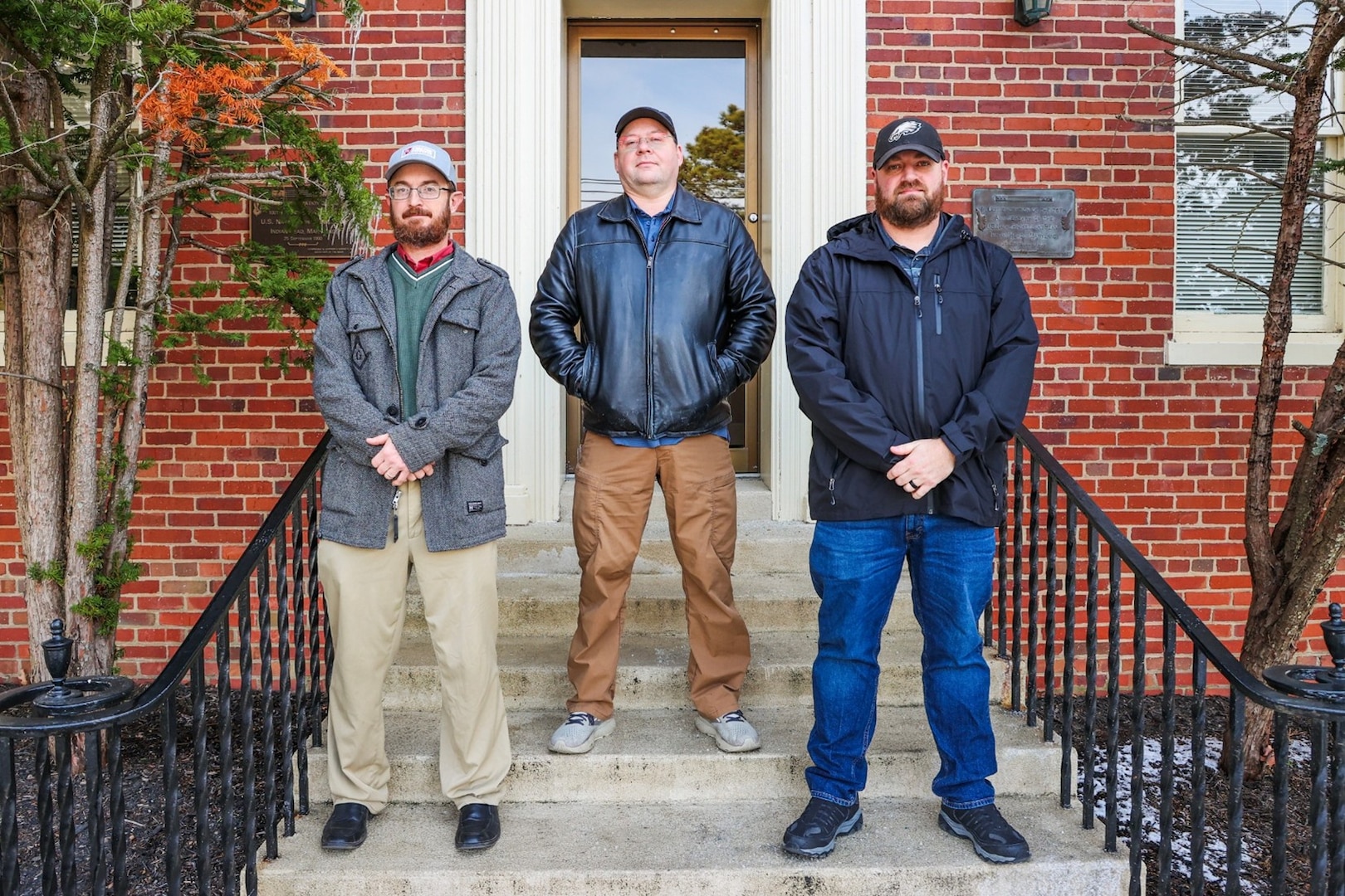 Photograph of three men from NSWC Indian Head's COMSEC team standing on steps in front of a brick building.