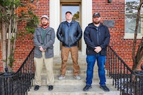 Photograph of three men from NSWC Indian Head's COMSEC team standing on steps in front of a brick building.
