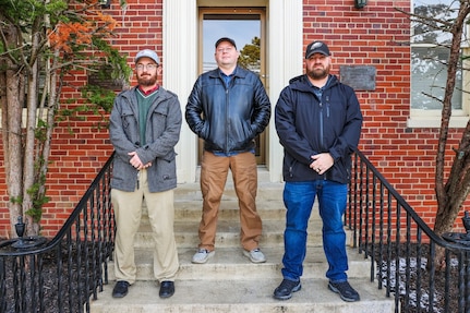 Photograph of three men from NSWC Indian Head's COMSEC team standing on steps in front of a brick building.