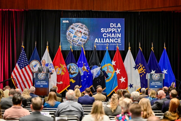 People in business attire fill a ballroom at a convention center to listen to a speaker. There are flags behind him as he stands at the podium.