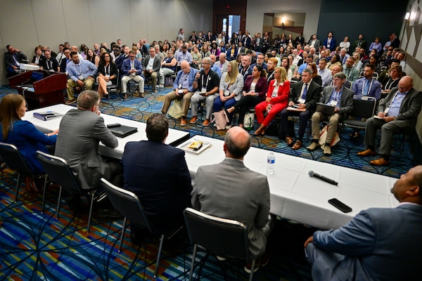 A multitude of people sit in a small room and listen to a panel of experts.