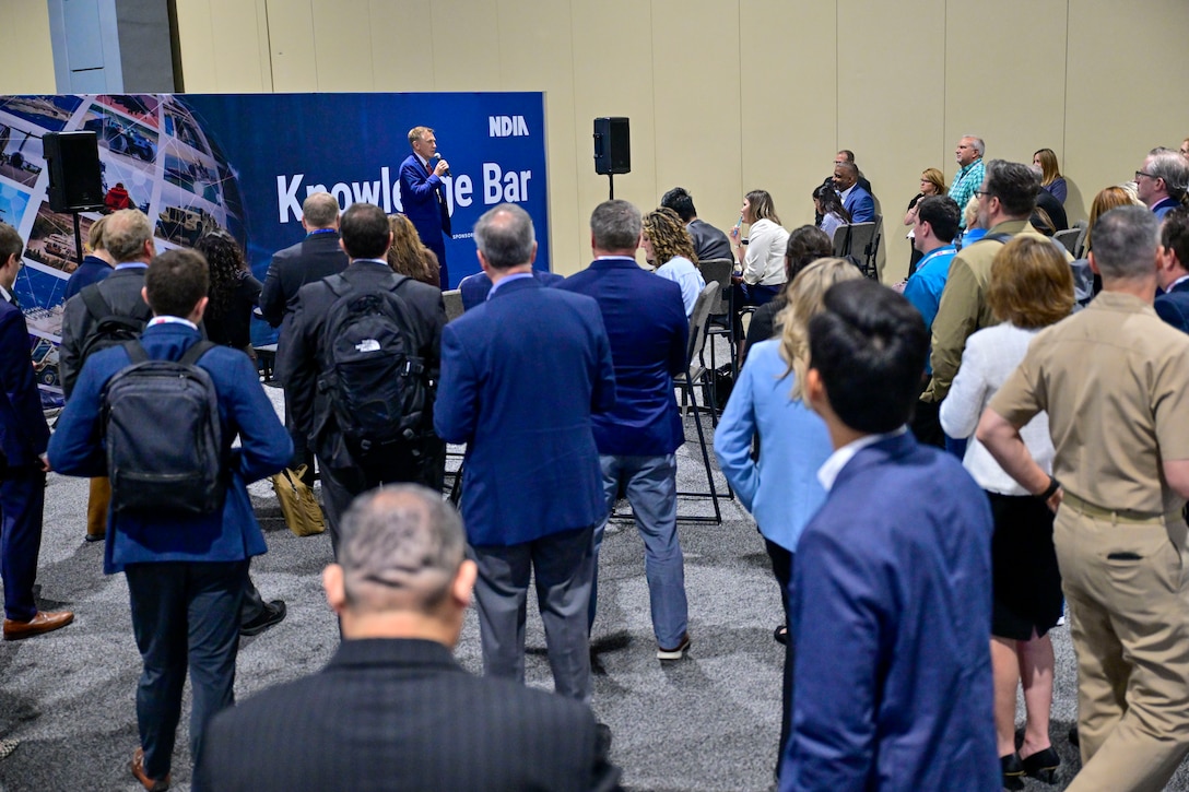 People in business attire listen to a speaker in a exhibition hall.
