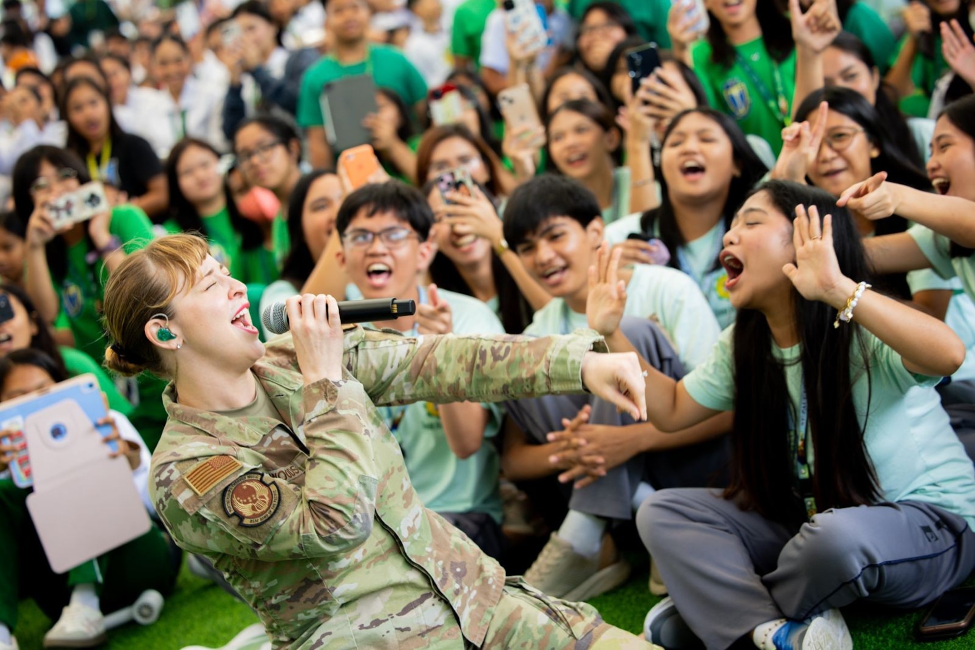 A female U.S. Soldier sings into a microphone in front a large group of students cheering.