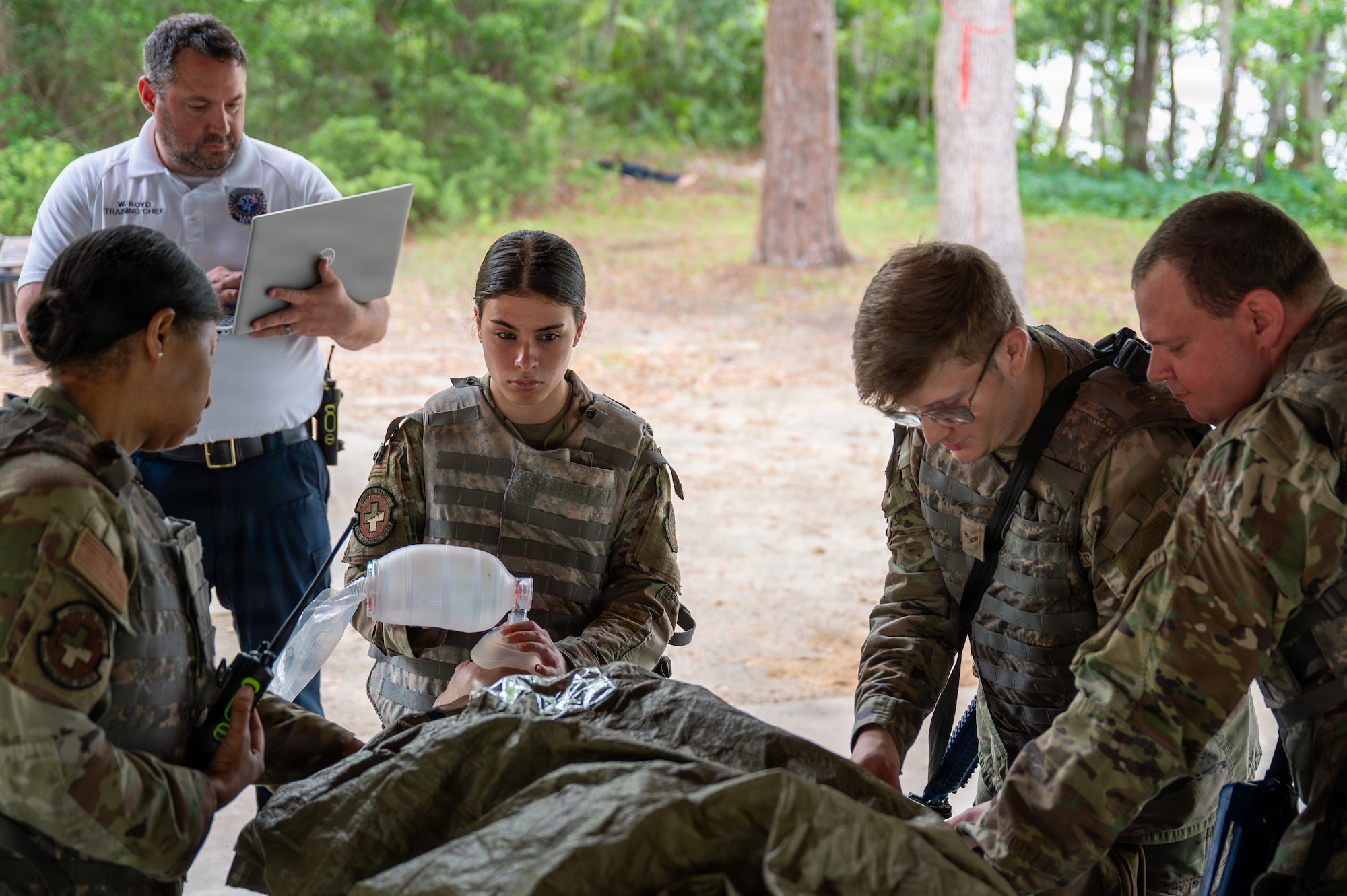 Four Airmen perform various medical tasks on a mannequin while a trainer stands behind them with a clipboard.