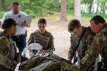 Four Airmen perform various medical tasks on a mannequin while a trainer stands behind them with a clipboard.