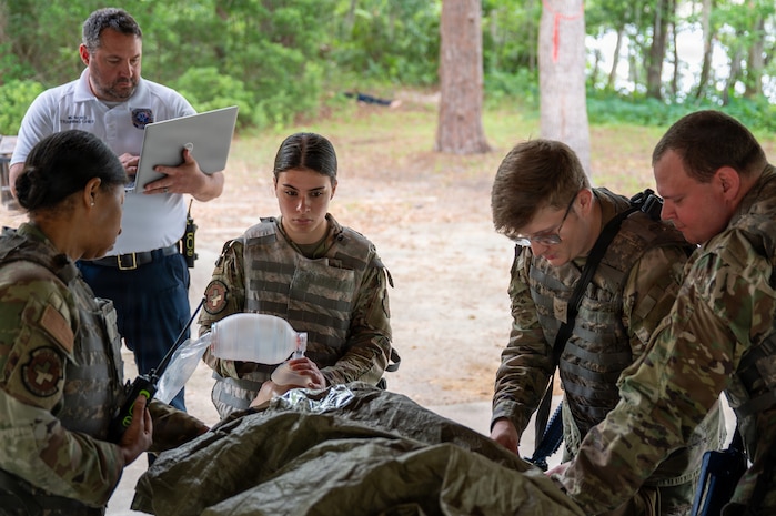 Four Airmen perform various medical tasks on a mannequin while a trainer stands behind them with a clipboard.