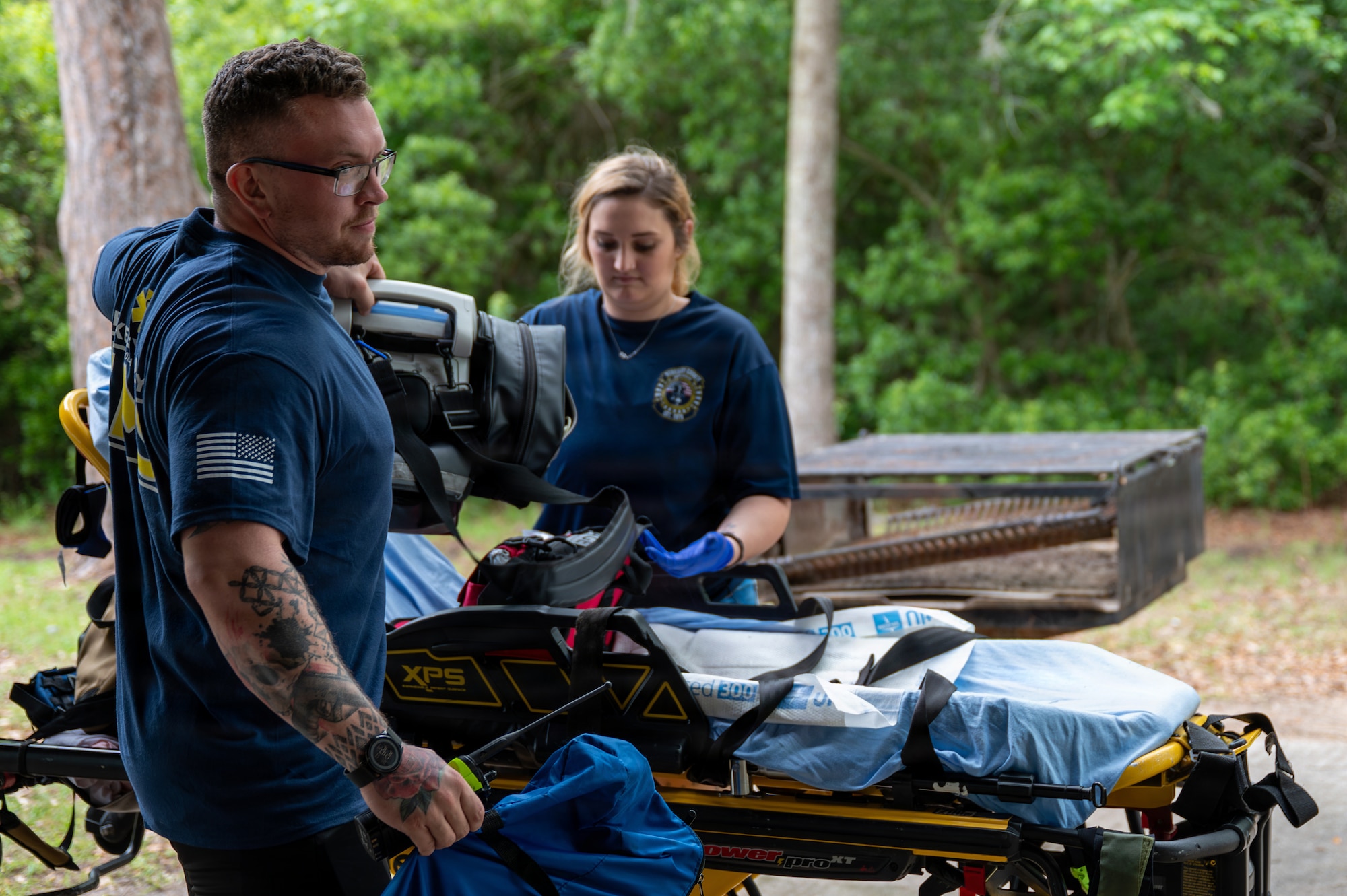 Two people attend to medical equipment standing next to a gurney.